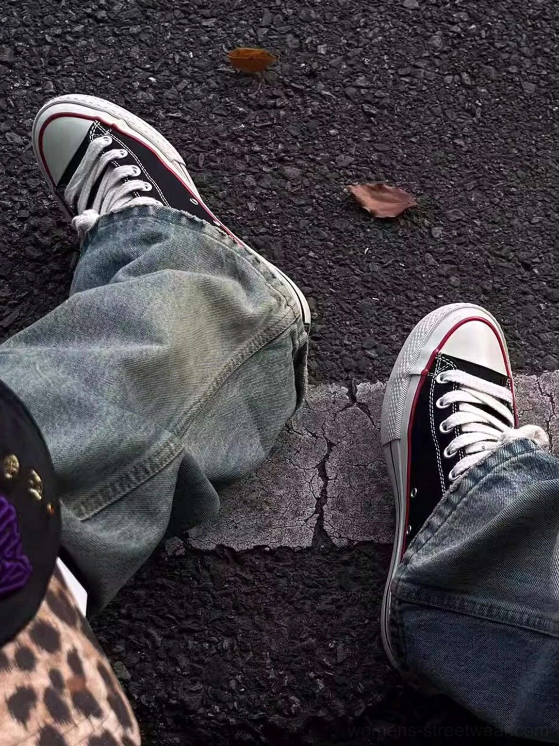 Black bat print canvas sneakers worn with loose blue denim jeans on an urban asphalt street with fallen leaves, showing red trim streetwear styling