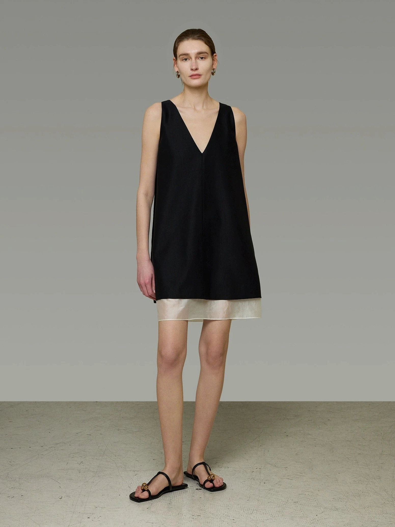 Model wearing a relaxed black and beige V-back shift dress with a loose A-line fit and black flat slide sandals in a minimalist studio