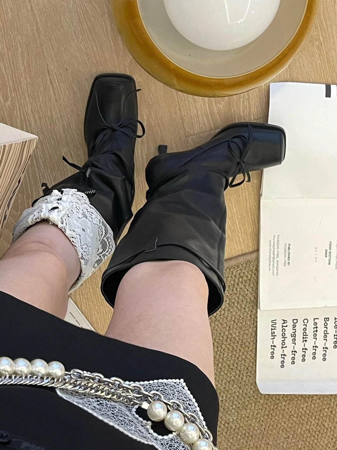Alternate overhead view of black bow knee-high leather boots with a softly gathered shaft and square toe styled in a feminine streetwear outfit