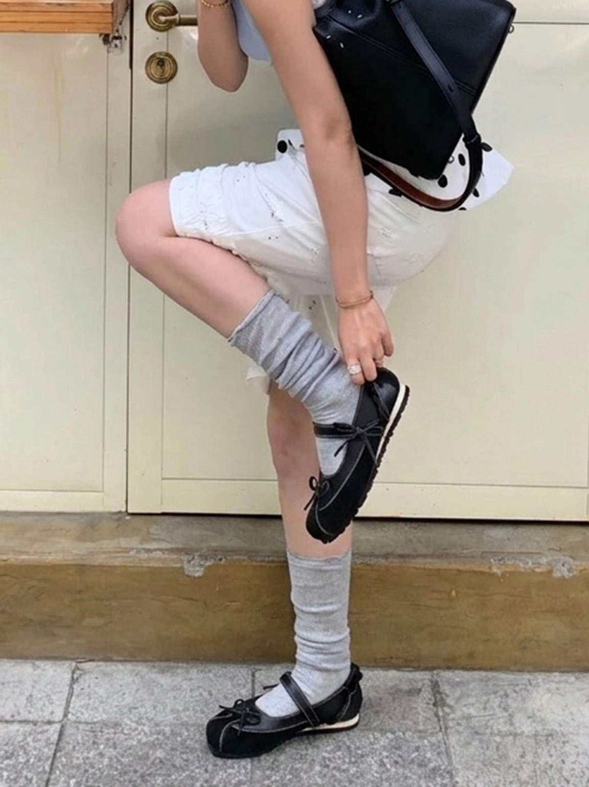 Black bow Mary Jane ballet flats worn with grey crew socks, white shorts and backpack in an outdoor streetwear setting