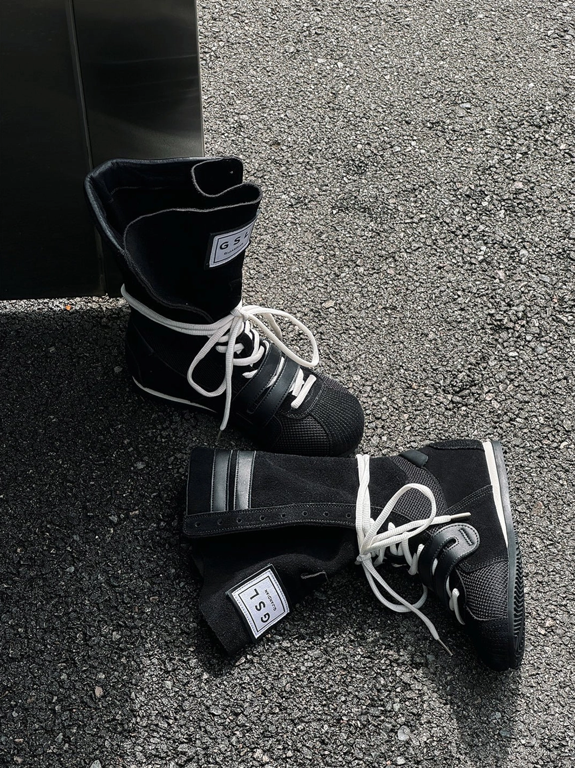 Overhead flat lay of black boxing trainer boots with white laces and square tongue tags on asphalt