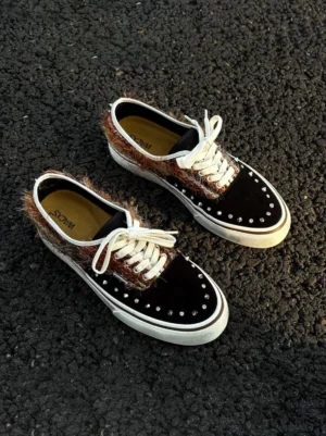 Black and brown mohair-panel studded low-top streetwear sneakers displayed overhead on asphalt, showing black suede toe caps, fuzzy contrast panels, silver rivets, white laces, and white rubber soles