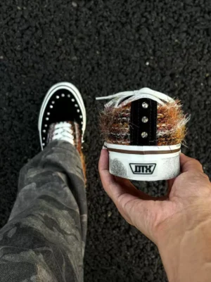 Close-up handheld view of black and brown studded low-top sneakers with a plush mohair-style heel collar, metal heel accents, layered suede textures, and detailed streetwear construction