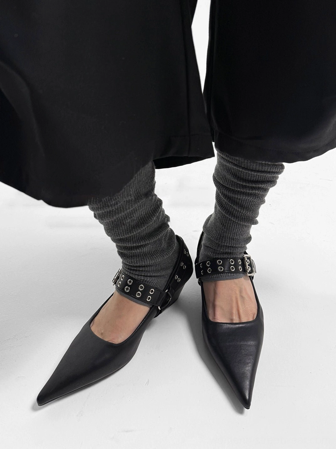 Pair of black buckle-strap pointed Mary Jane heels worn with charcoal leg warmers and modern dark streetwear