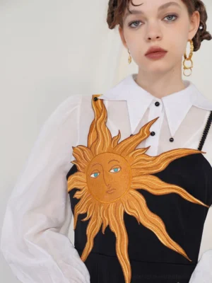 Close-up view of golden sun face embroidery with blue eyes on a black camisole maxi dress styled over a sheer white collared blouse