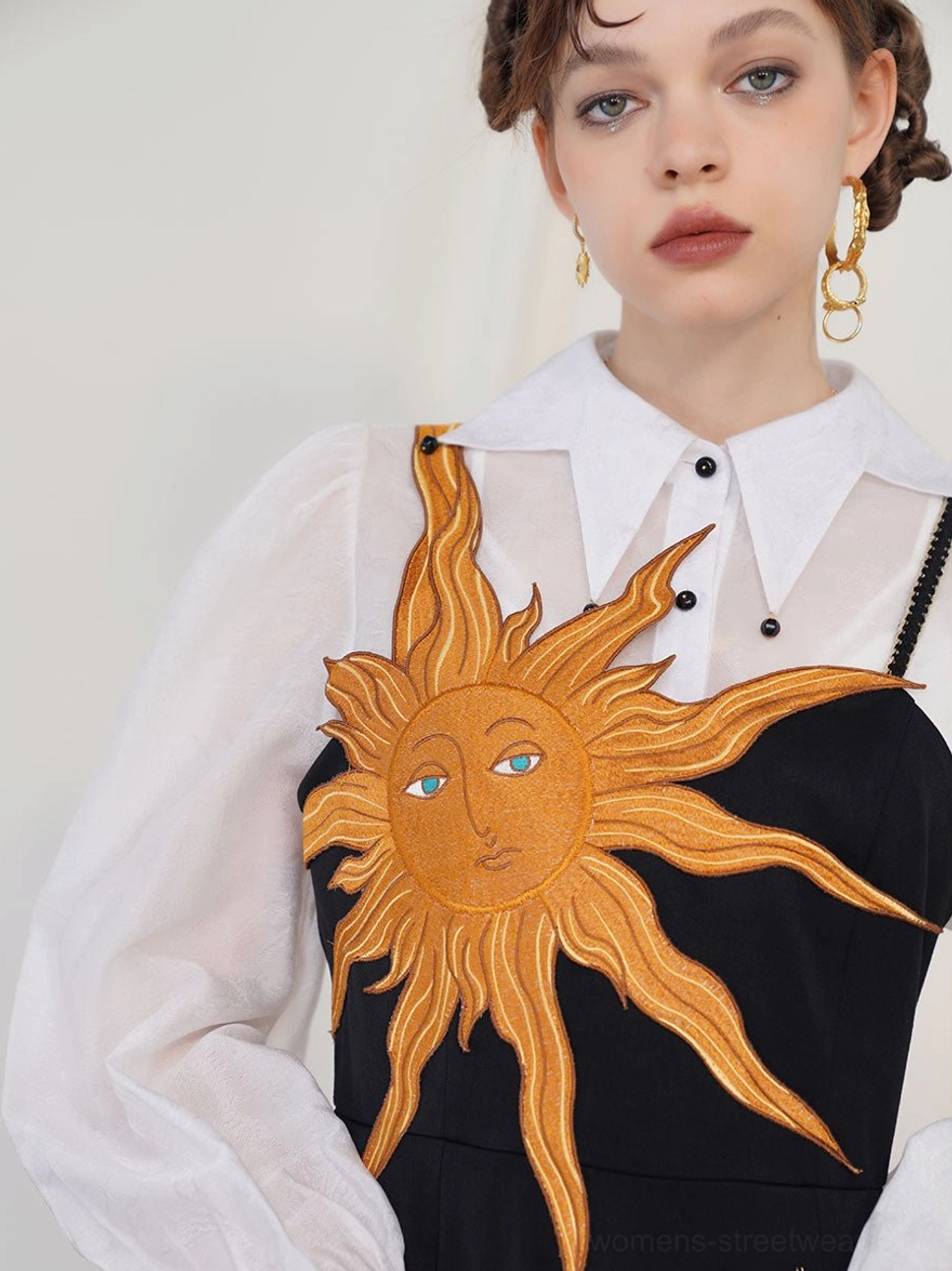 Close-up view of golden sun face embroidery with blue eyes on a black camisole maxi dress styled over a sheer white collared blouse