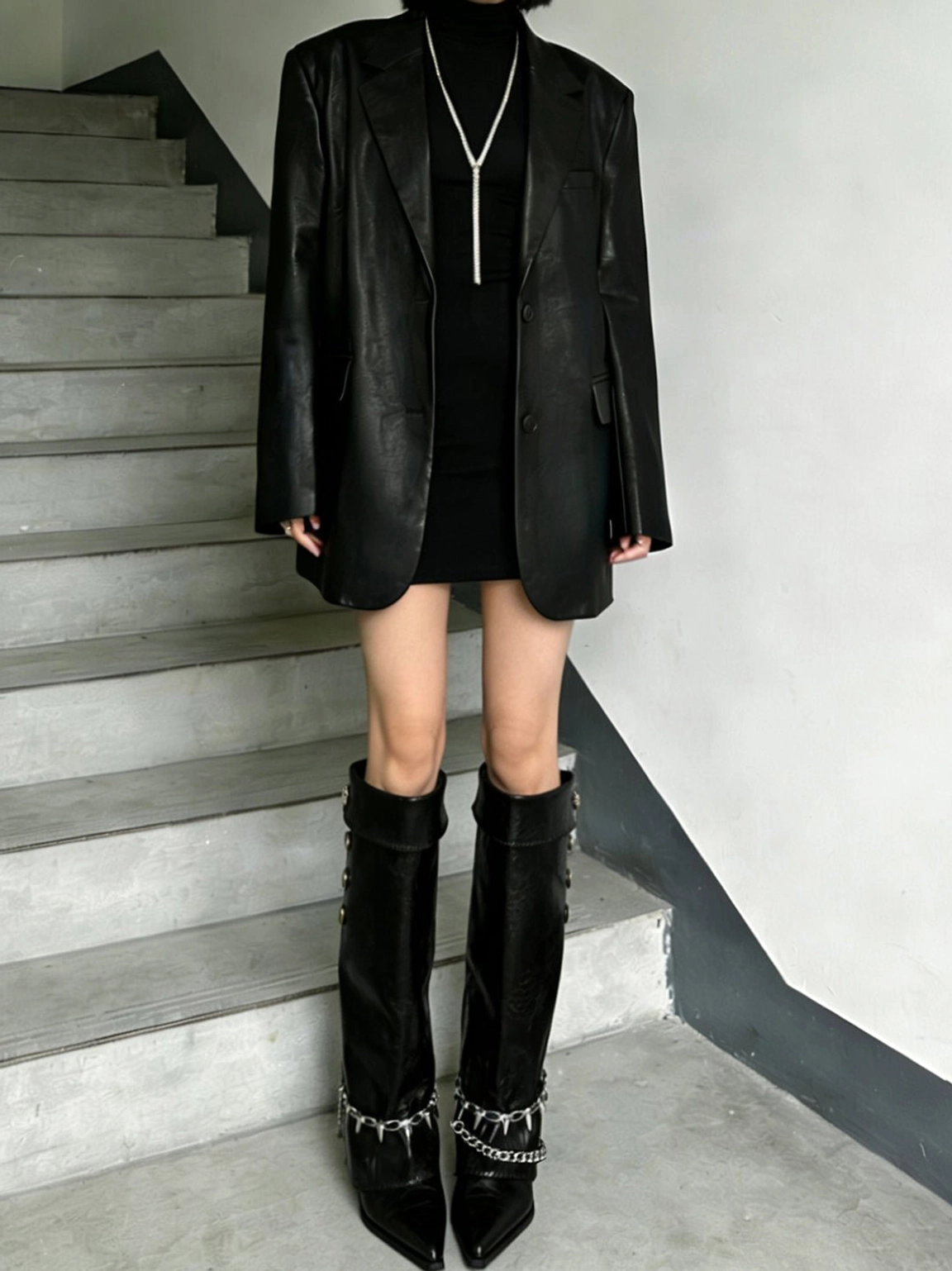 Front view of black chain-trim pointed riding boots styled with a black blazer and mini dress, showing layered ankle chains and pointed flat toes