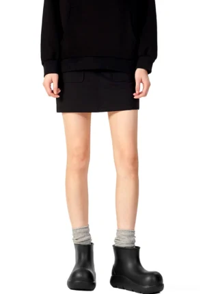 Model styling black chunky ankle rain boots with grey crew socks, mini skirt, and cropped black sweatshirt in a streetwear outfit