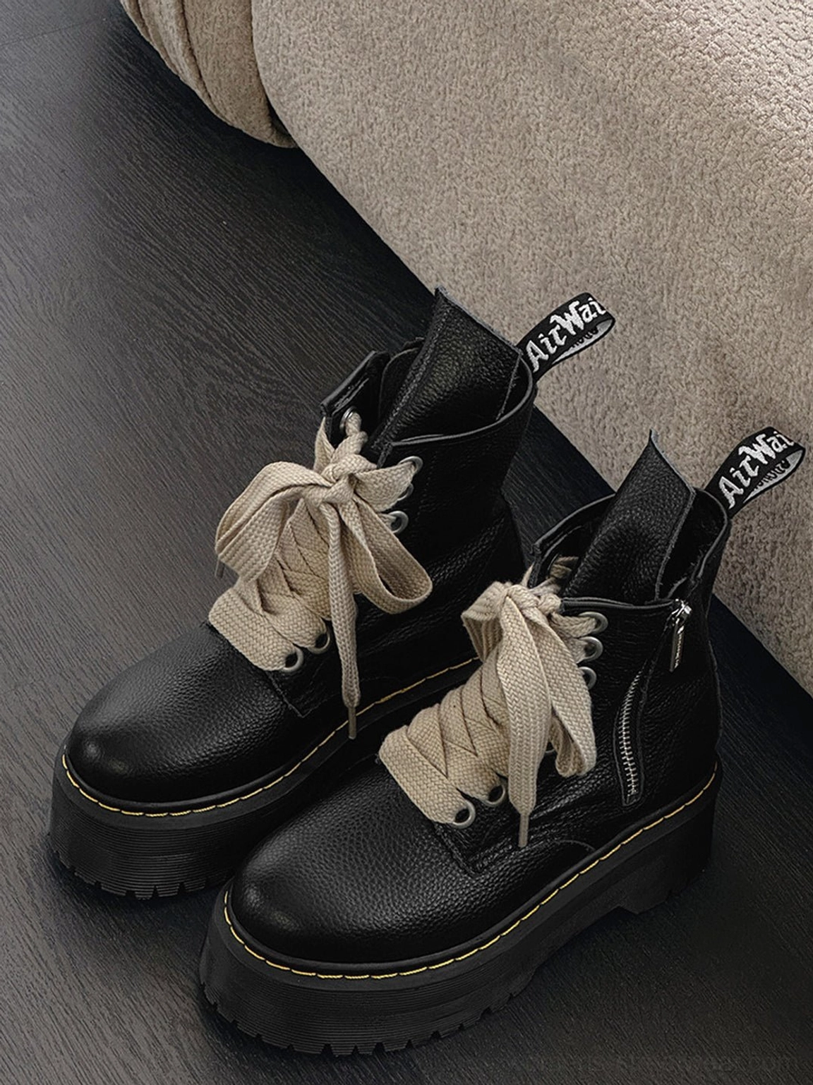 Flat lay of black chunky platform combat boots with wide cream ribbon laces, yellow sole stitching, branded zipper pulls, and side closures