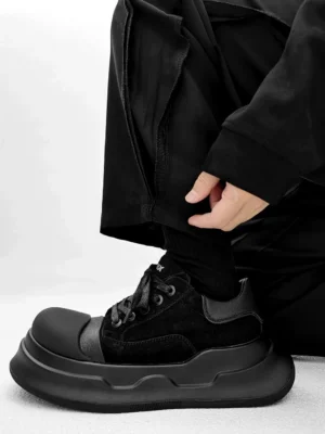 Side profile of black chunky platform sneakers worn with loose cargo pants, featuring a thick layered outsole, rounded toe cap, padded collar, and monochrome streetwear design
