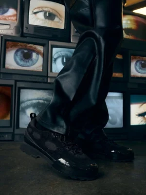 Black chunky streetwear sneakers worn with black leather pants in front of retro televisions, showing the oval cutout upper and thick rubber outsole