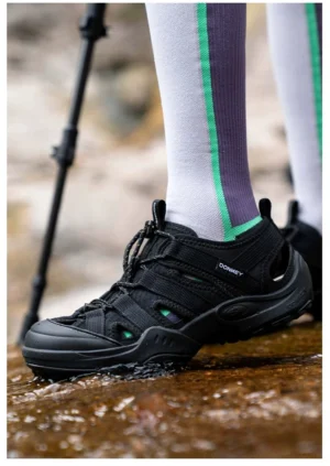 Black closed-toe outdoor water sandals worn with green striped knee-high socks while walking through shallow rocky water, showing breathable upper and secure ankle fit