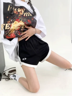 Front kneeling view of black contrast trim dolphin hem track shorts with white piping, elastic waistband, and circular embroidered emblem in a minimalist studio