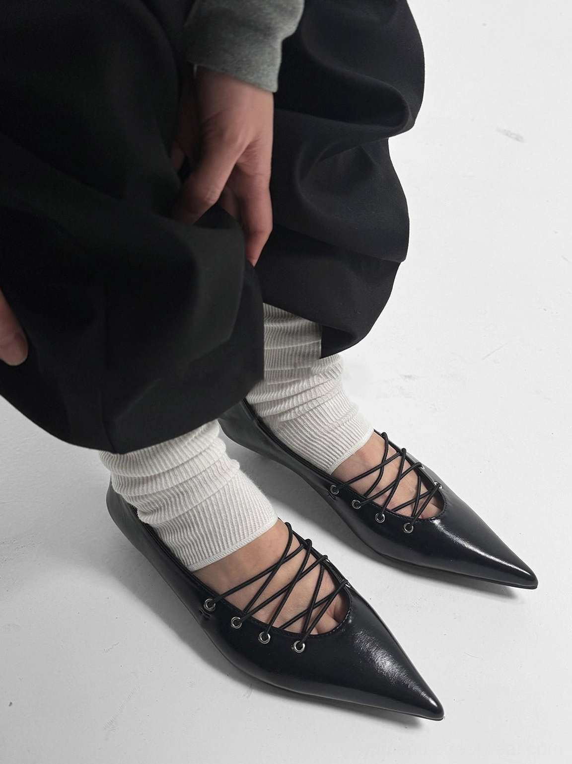 Black corset-lace pointed ballet flats worn with white slouch socks, loose trousers, and a gray long-sleeve top