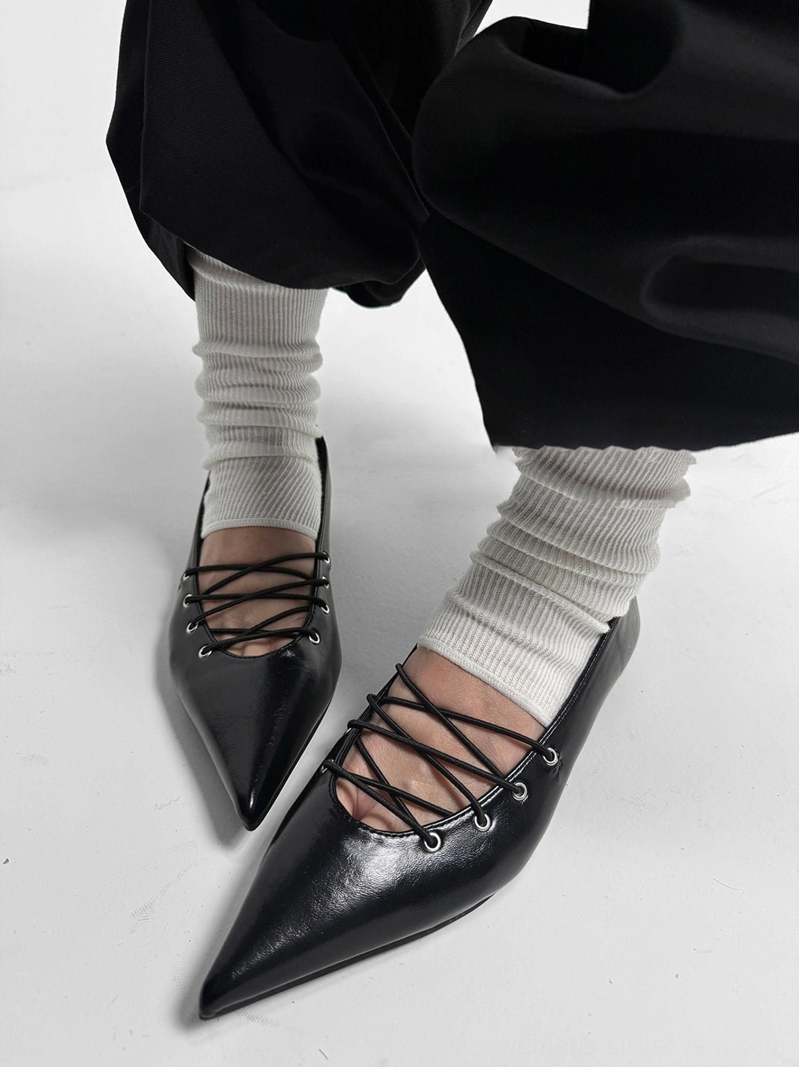 Top view of black corset-lace pointed ballet flats worn with white slouch socks and wide-leg black trousers