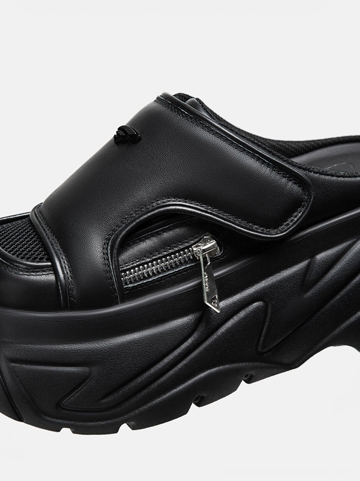 Close-up angled view of black covered-toe platform slide sandal with thick textured outsole, adjustable strap, silver zipper, and matte streetwear finish