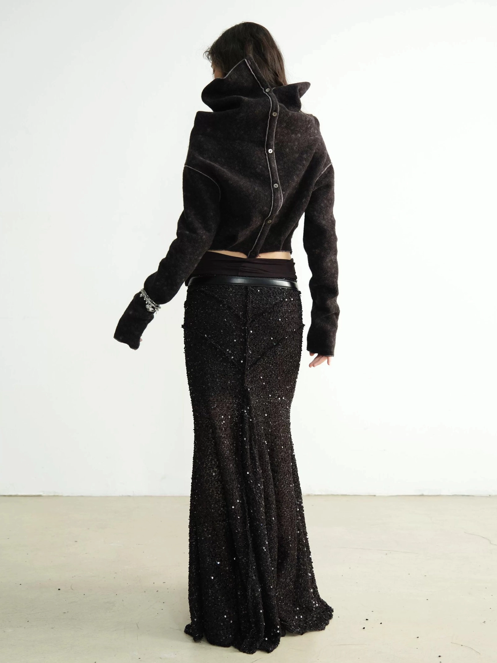 Back view of a black cowl-neck cropped knit cardigan with exposed center seam, extended cuffs, and high-waisted mermaid skirt