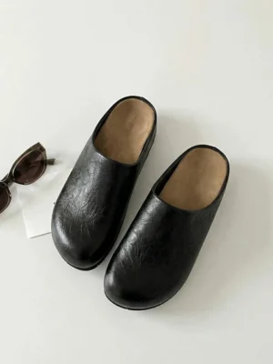 Pair of black cracked leather closed-toe mule clogs shown from above with tan insoles, sunglasses, and minimalist streetwear styling