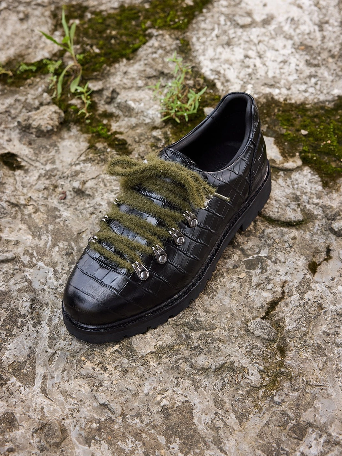 Three-quarter view of a black croc-embossed leather derby shoe with olive laces, rounded toe, metal eyelets, and lug outsole
