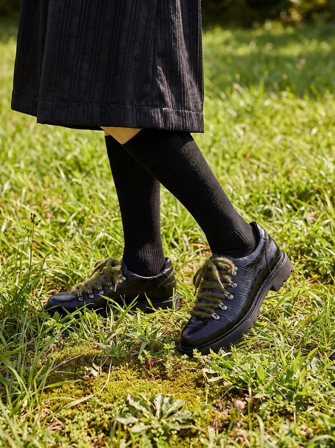 Black croc-embossed leather derby shoes styled with black pleated skirt and knee-high socks in an outdoor streetwear look