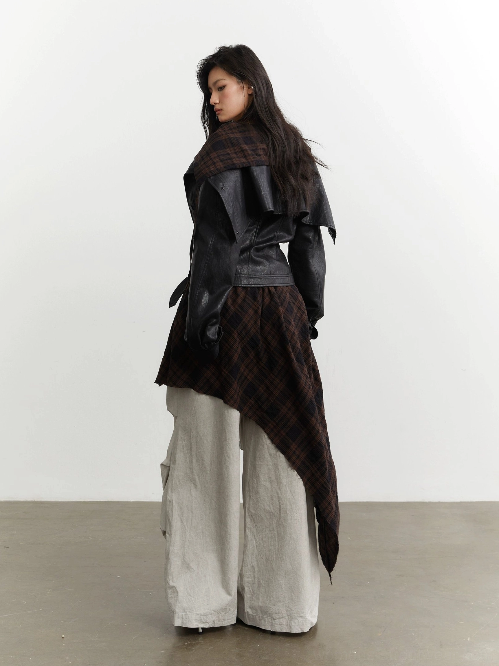 Side back view of a black cropped faux leather jacket with flowing asymmetric tartan plaid hem layered over wide-leg cargo pants