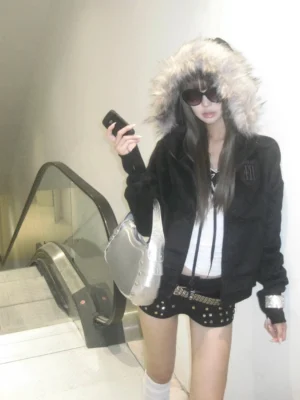 Full-body model wearing a black cropped fur hooded zip hoodie with ribbed cuffs, subtle chest embroidery, white crop top, and studded mini shorts
