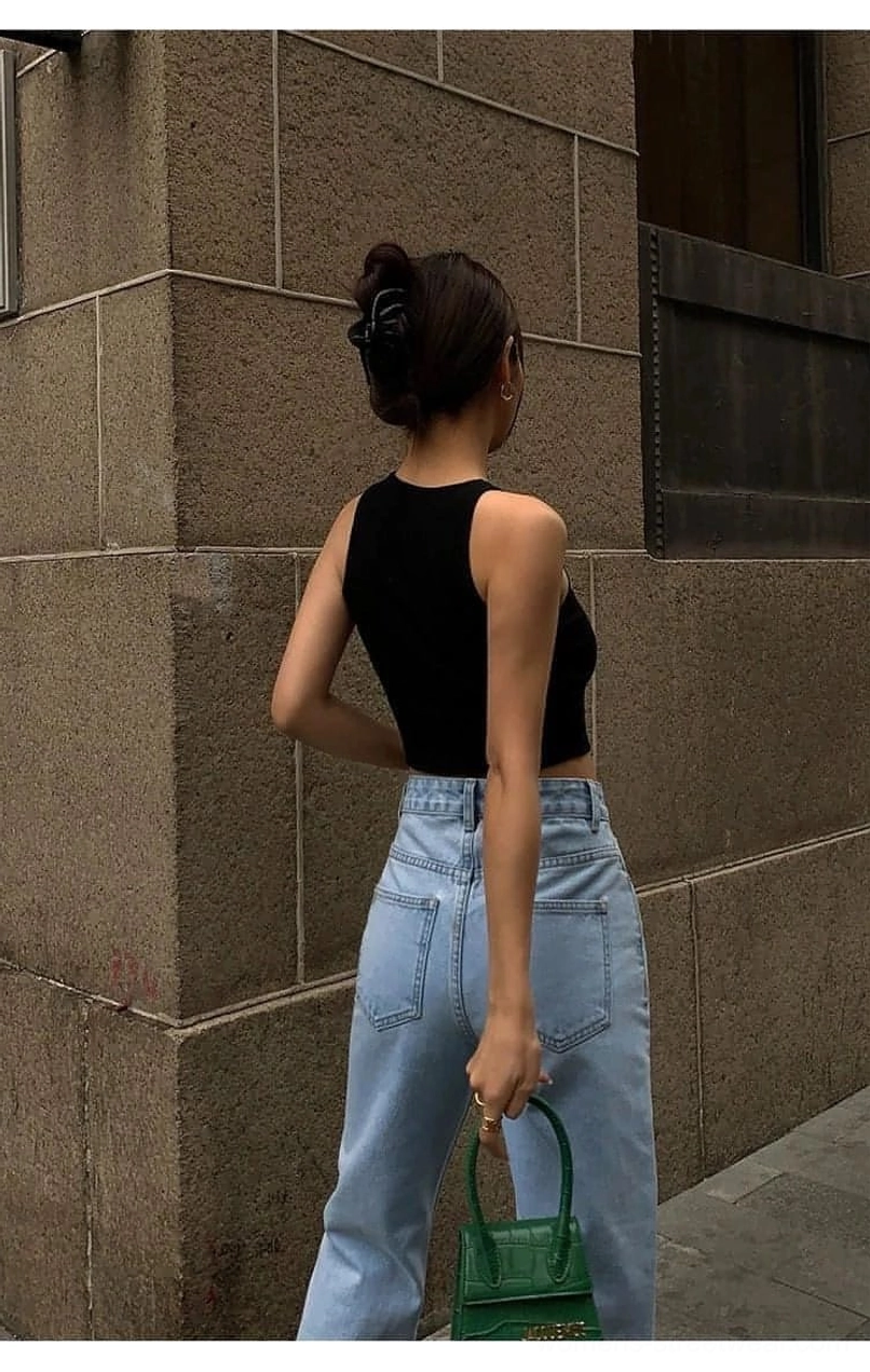 Black cropped knitted tank top back close-up shot - slim cropped silhouette showing exposed back waist and ribbed knitted fabric texture paired with light blue jeans and green handbag on stone wall outdoor setting