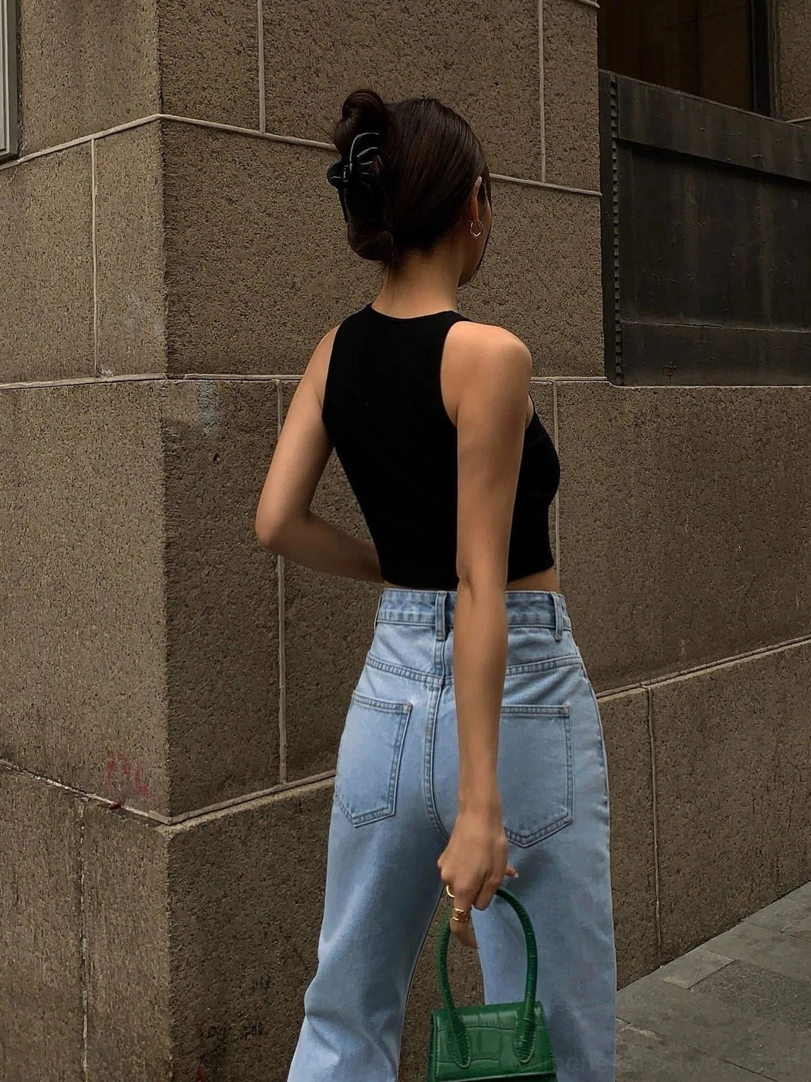 Black cropped knitted tank top back view - sleeveless design showing exposed waistline and ribbed knit texture paired with high-waisted light blue jeans and green handbag on stone wall outdoor setting