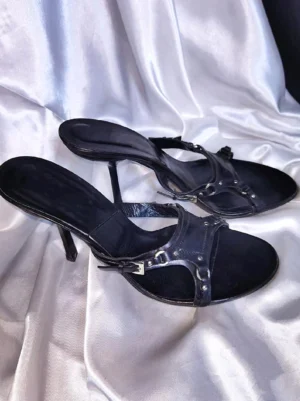 Black open-toe stiletto sandals with crossed vamp straps and metallic buckle details shown from an angled side view on silver satin fabric