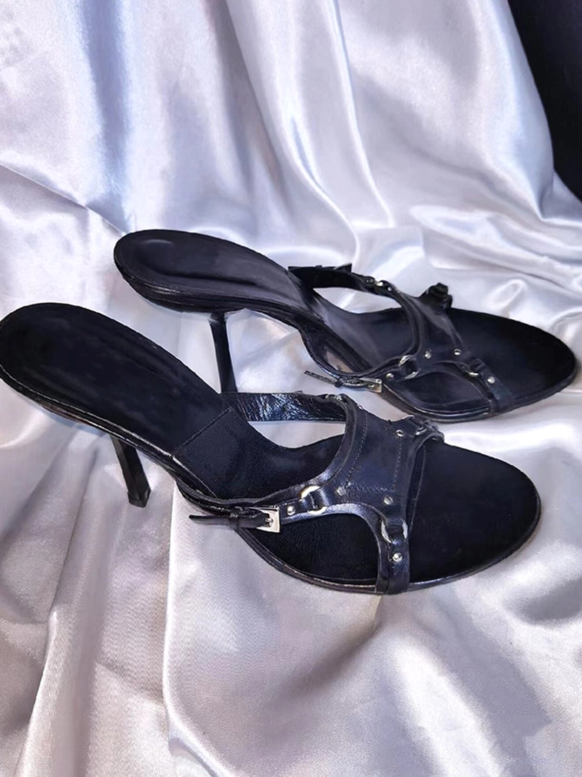 Black open-toe stiletto sandals with crossed vamp straps and metallic buckle details shown from an angled side view on silver satin fabric