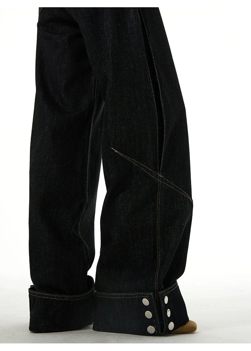 Close-up of black cuffed denim jeans showing multiple metal button fasteners, rolled hem, and diagonal decorative seam