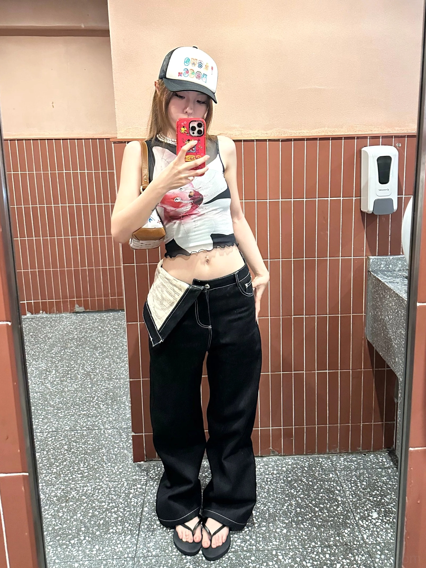 Mirror selfie showing black deconstructed waistband jeans styled with a printed crop top and baseball cap