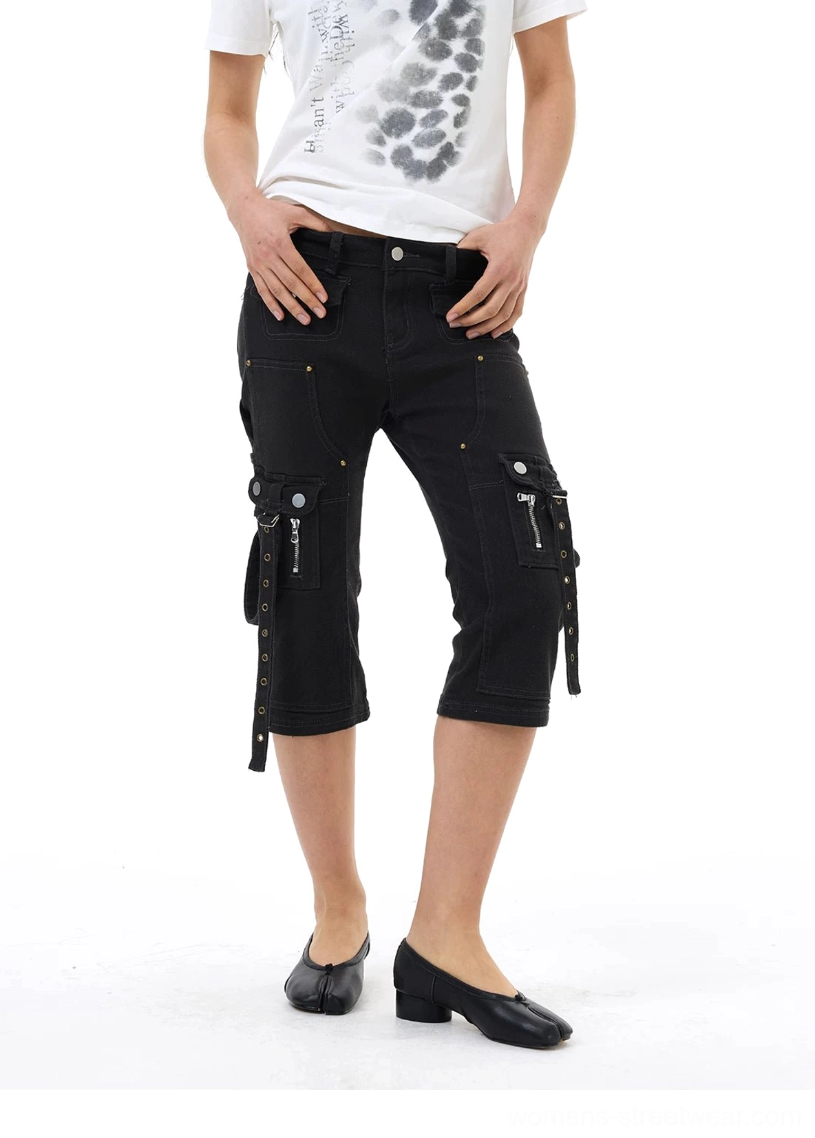 Close front styling view of black denim cargo capri jeans with metal button closure, dual zip pockets, hanging straps, and rivet hardware