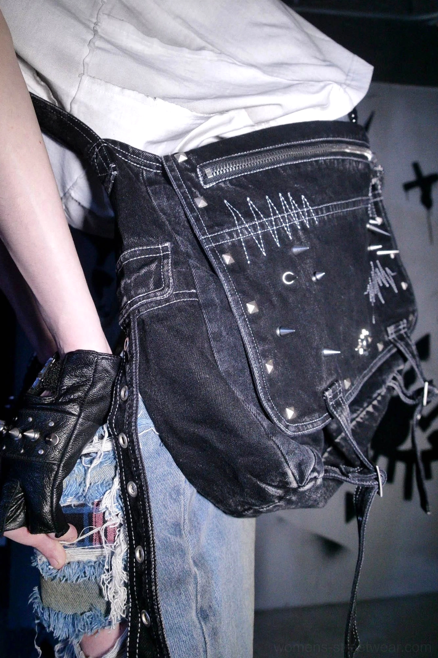 Handheld close-up of black washed denim cyberpunk utility bag showing zippered flap pocket heartbeat stitching metal rivets and charms