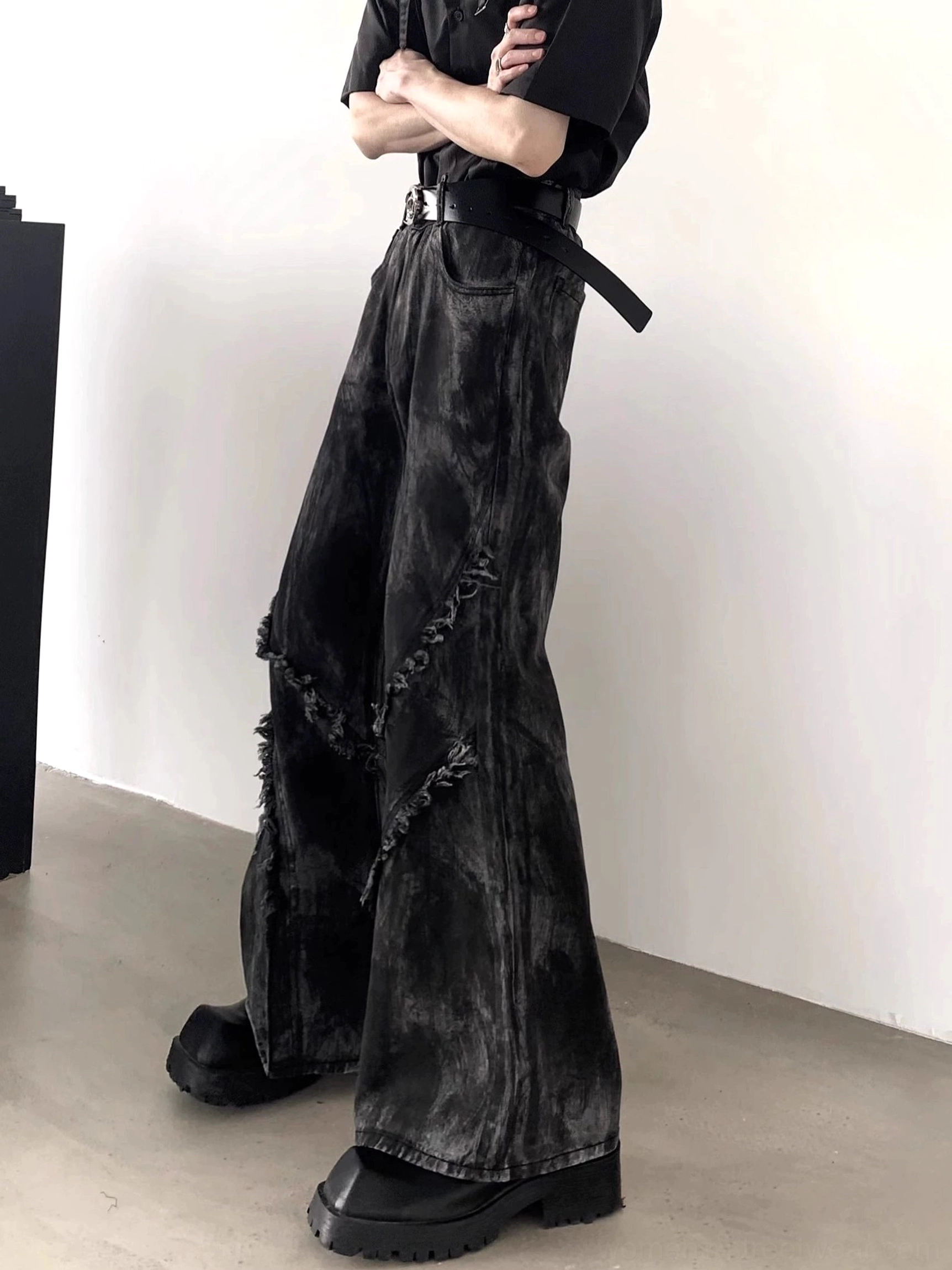 Side view of black distressed flare jeans featuring diagonal raw frayed seams, tie-dye washed denim, and an exaggerated wide-leg cut