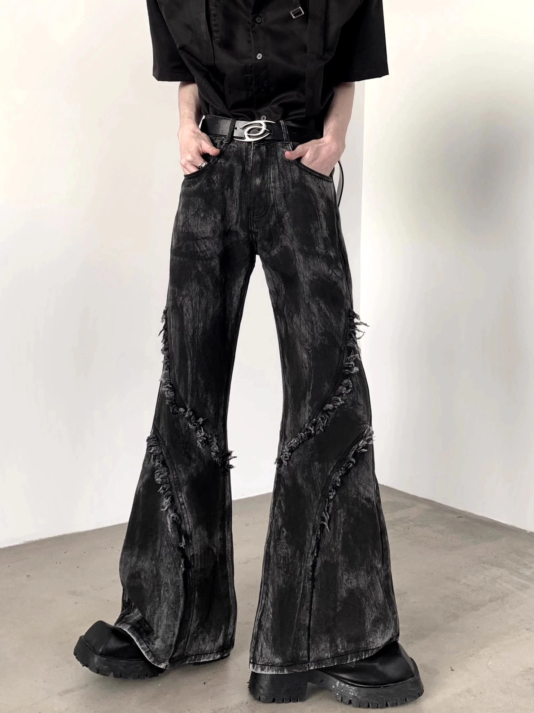 Front low-angle view of black distressed flared jeans with diagonal frayed seams, washed denim finish, wide-leg silhouette, belt, and platform boots