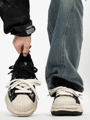 Close-up of black distressed shell-toe sneakers with dual flat laces, contrasting off-white toe reinforcement, textured fabric upper, and exaggerated wavy rubber sole