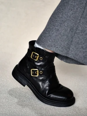 Black double buckle lambskin ankle boots worn with grey wide-leg trousers in a side three-quarter streetwear styling view