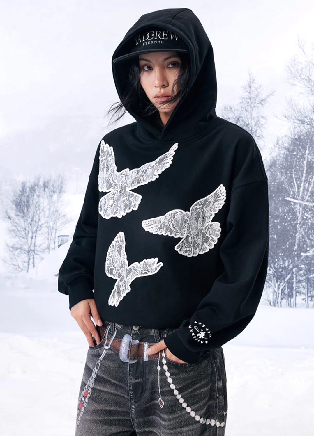 Front half-body model view wearing a black dove appliqué hoodie with vintage jeans, chain waist detail and studded accessories