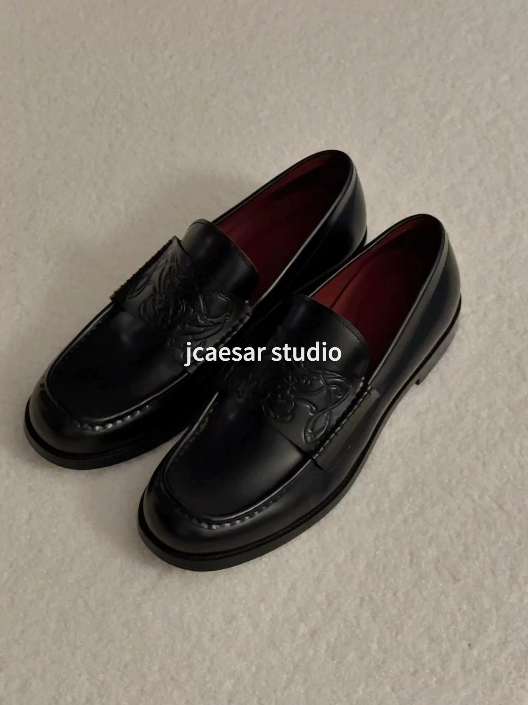 Pair of black embossed leather loafers displayed on a light beige fabric background with glossy leather, burgundy lining, and decorative vamp patterns
