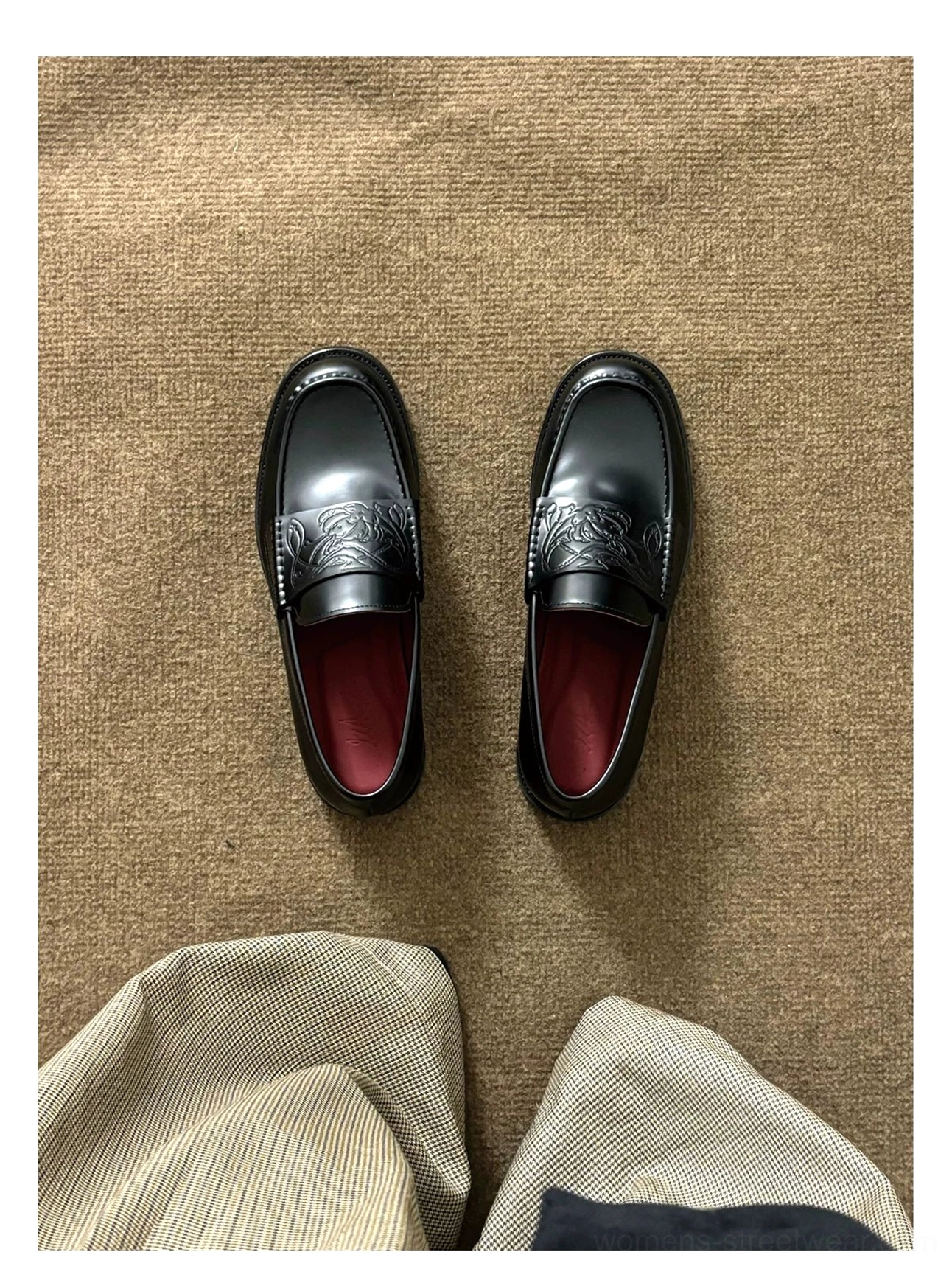 Pair of black embossed leather loafers on a brown woven fabric background with glossy leather, burgundy lining, and matching trouser hems
