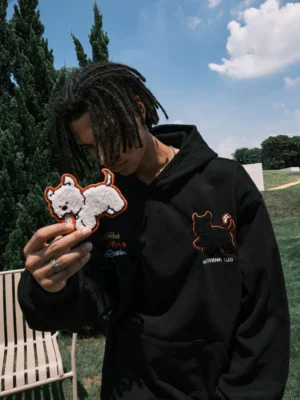 Black oversized embroidered dog pullover hoodie shown from the front on a streetwear model outdoors, featuring textured puppy chest embroidery, script lettering, kangaroo pocket, and matching dog patch