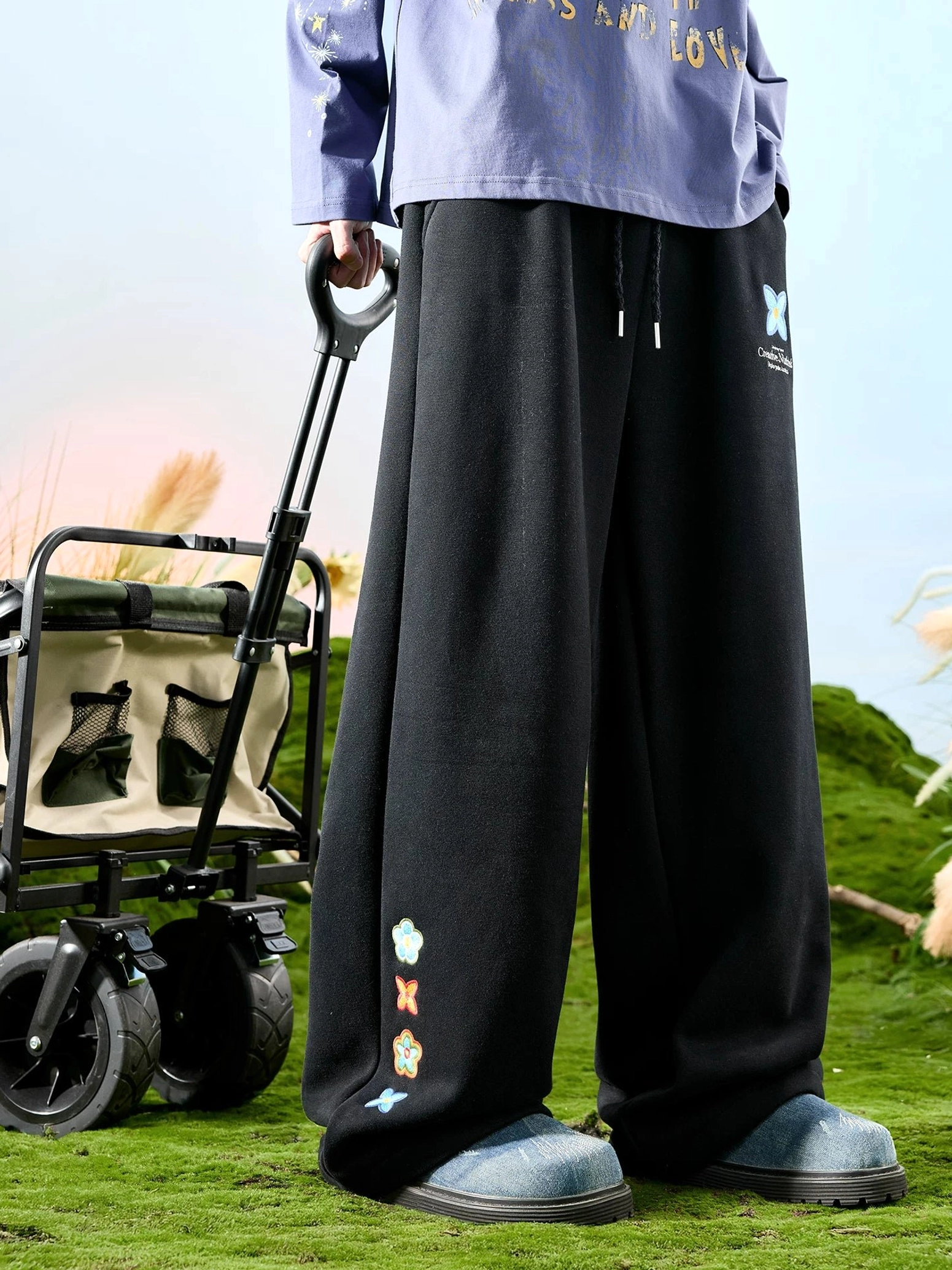 Black embroidered floral wide-leg streetwear sweatpants with blue butterfly detail worn in an outdoor front view