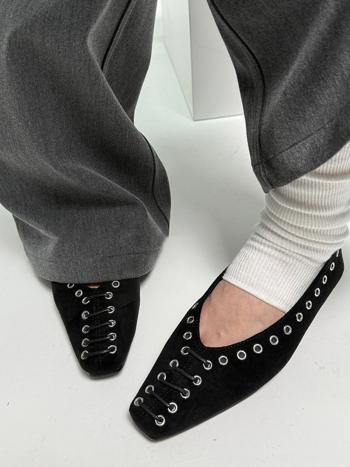 Side view of black eyelet ballet flats worn with grey cropped trousers and white slouch socks