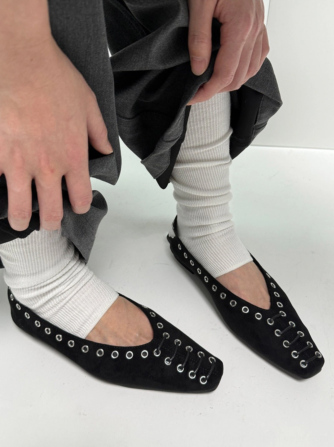 Pair of black eyelet-trim square-toe ballet flats styled with grey loose trousers and white slouch socks