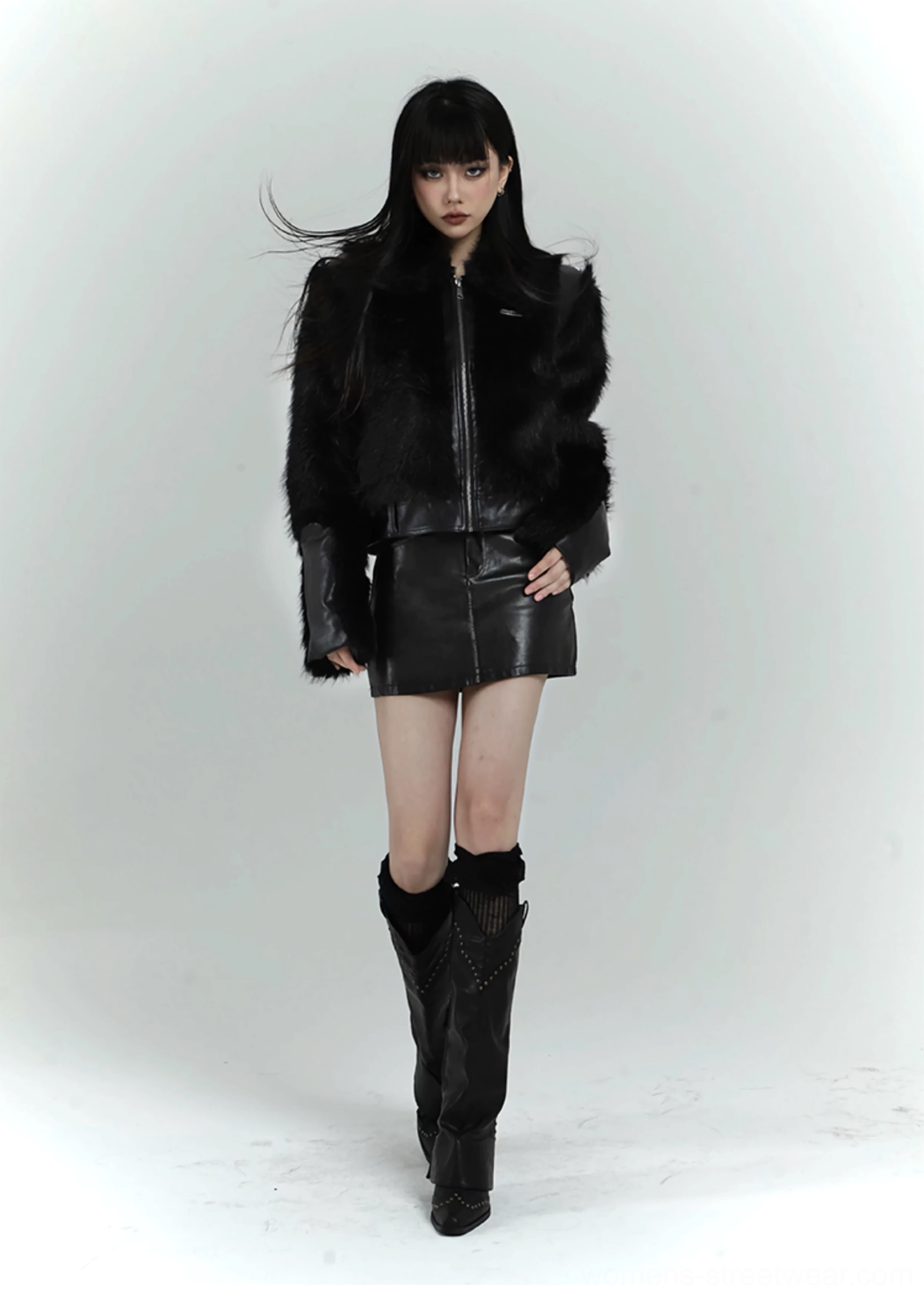Low-angle view of a black faux fur cropped bomber jacket styled with a leather-look skirt and studded boots