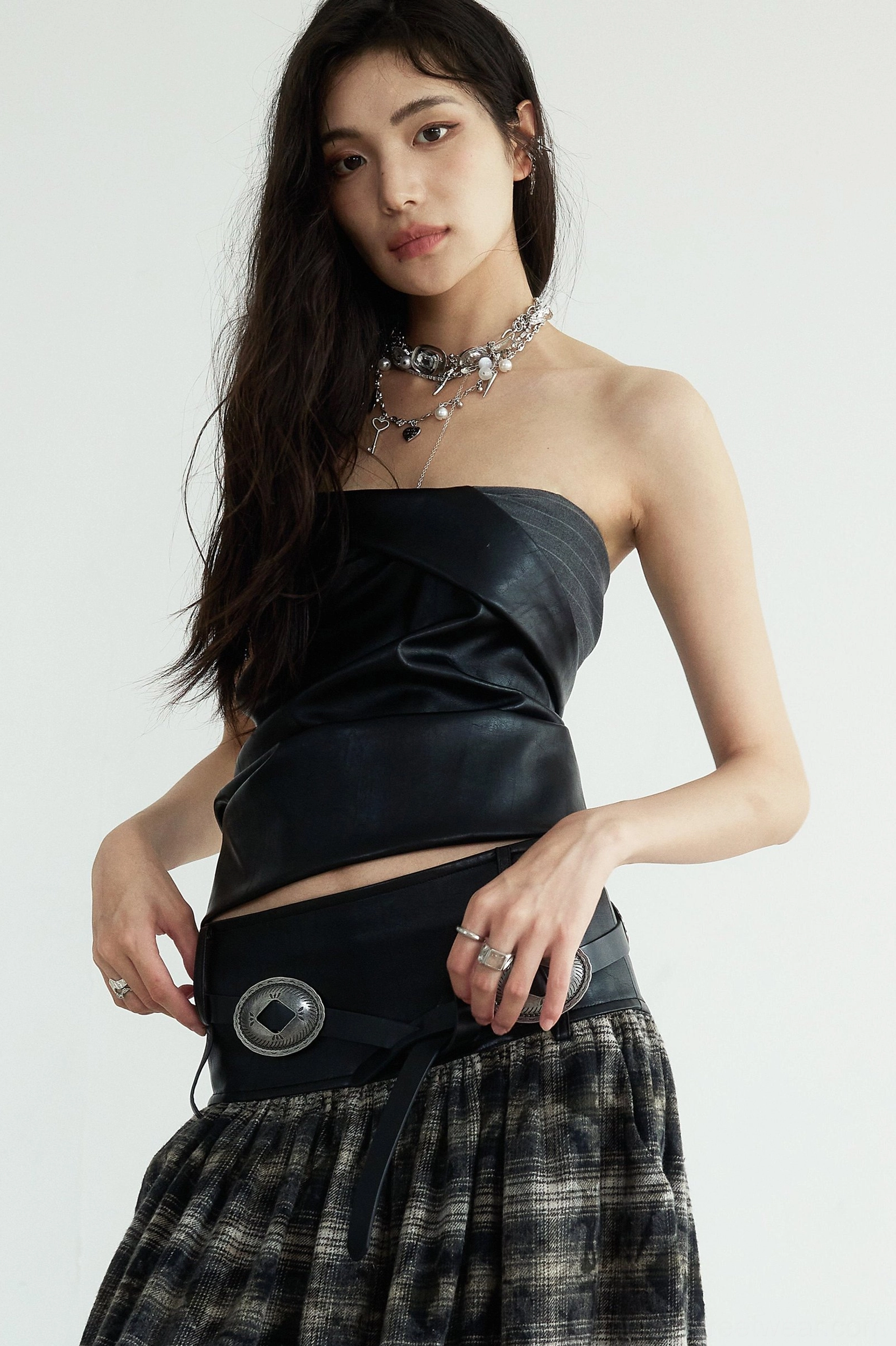Front view of a black faux leather origami bustier top with strapless corset silhouette, cropped waist, metal buckle detail, and plaid maxi skirt styling