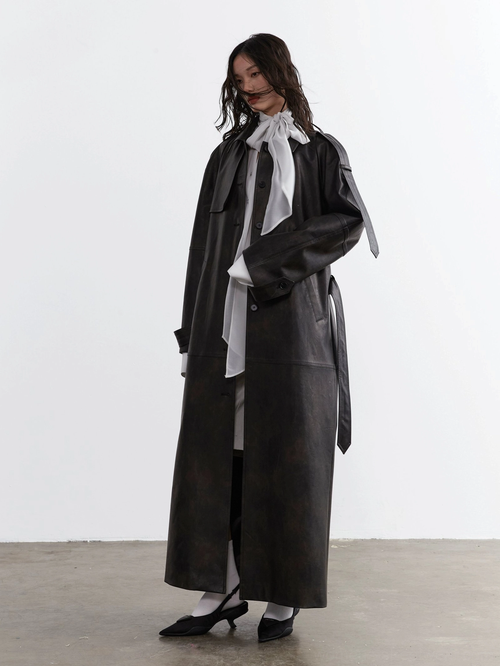 Black faux leather trench coat styled over a sheer long-sleeve neck-tie streetwear blouse