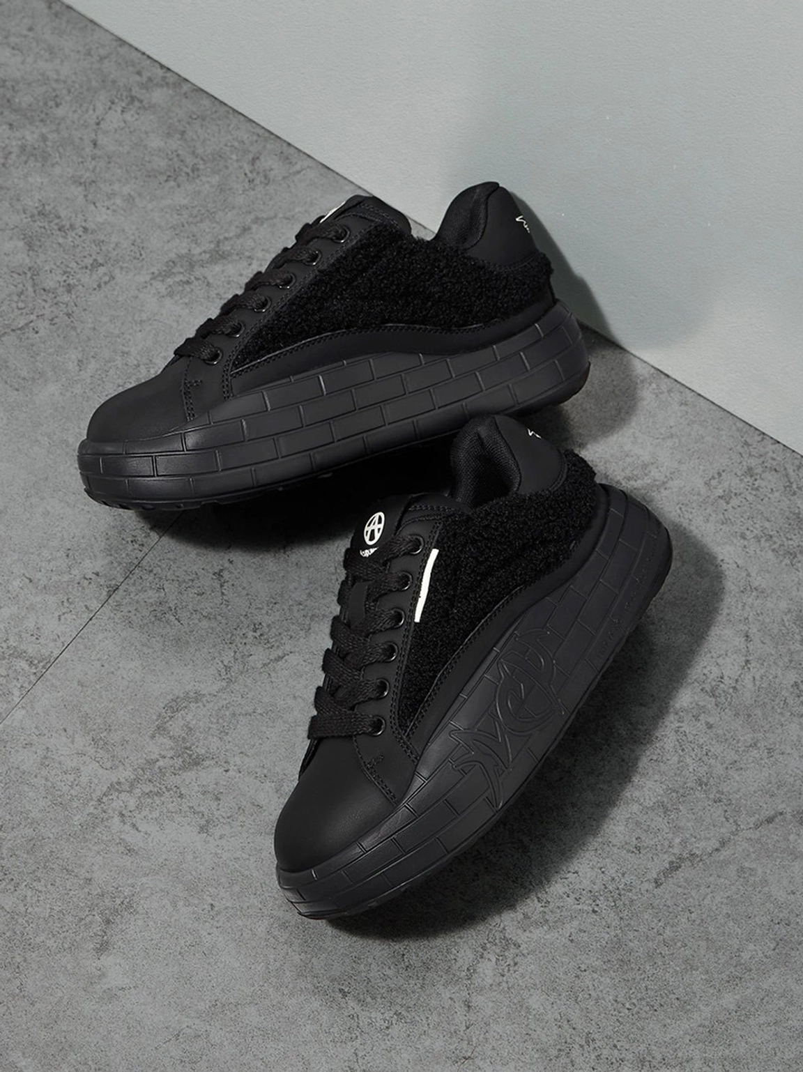 Black fleece platform sneakers shown separately on a concrete floor with matte toe caps and textured soles