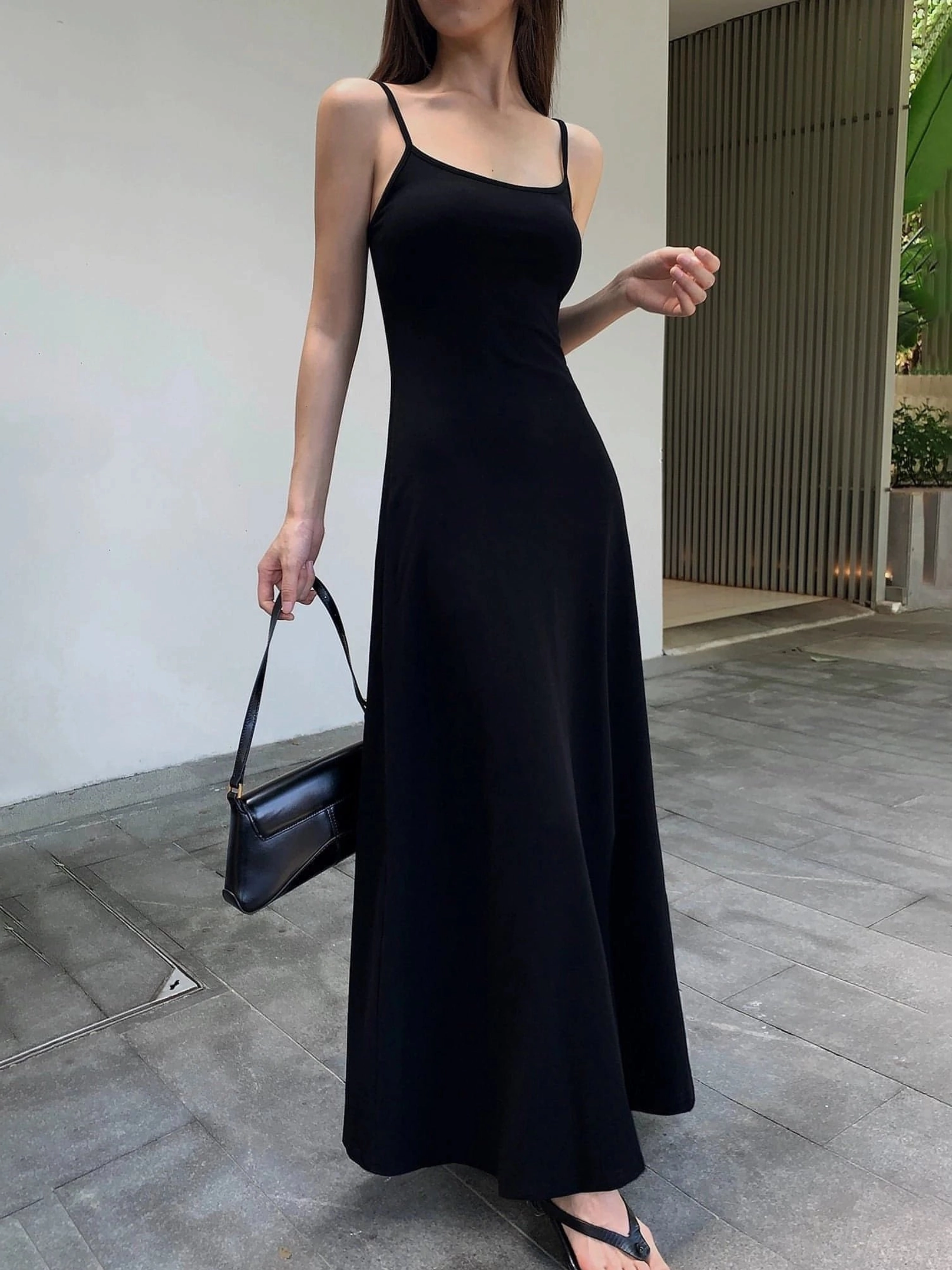 Black floor length spaghetti strap slip dress styled with a black leather underarm bag in a modern outdoor setting